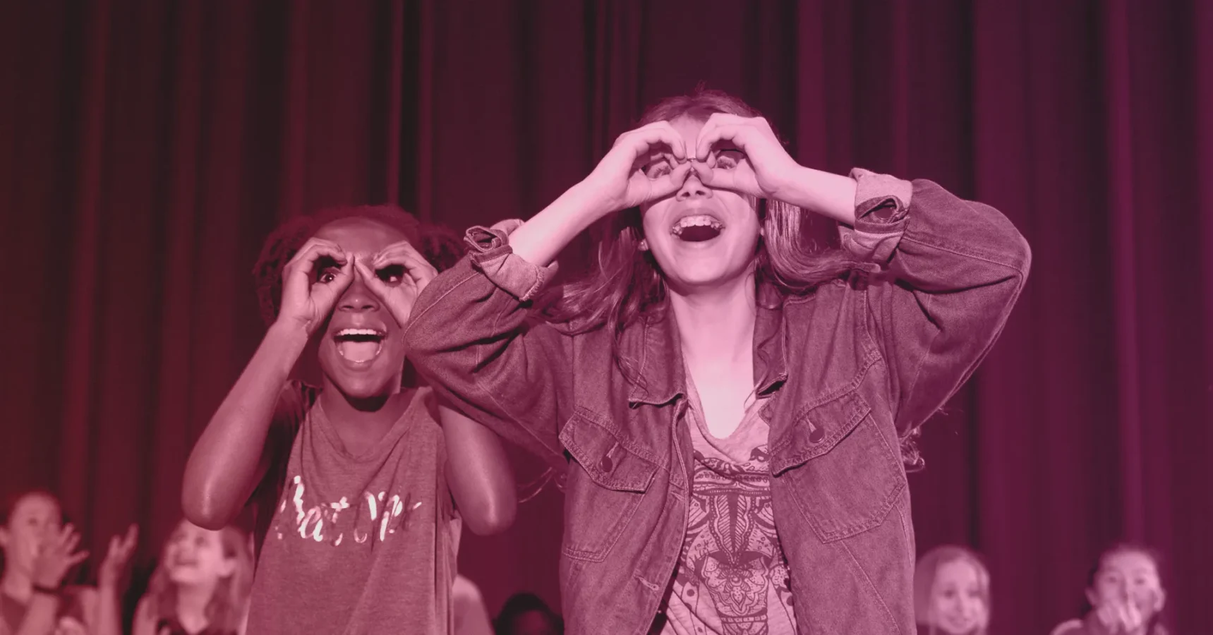 Two children stand side by side, smiling and making pretend binoculars with their hands in front of a dark curtain backdrop.