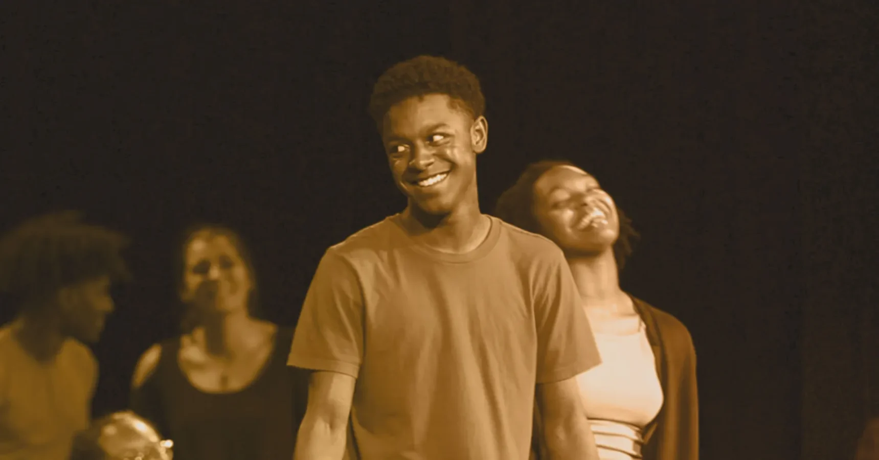 A young man and woman stand on stage smiling, with two other people blurred in the background against a dark backdrop.