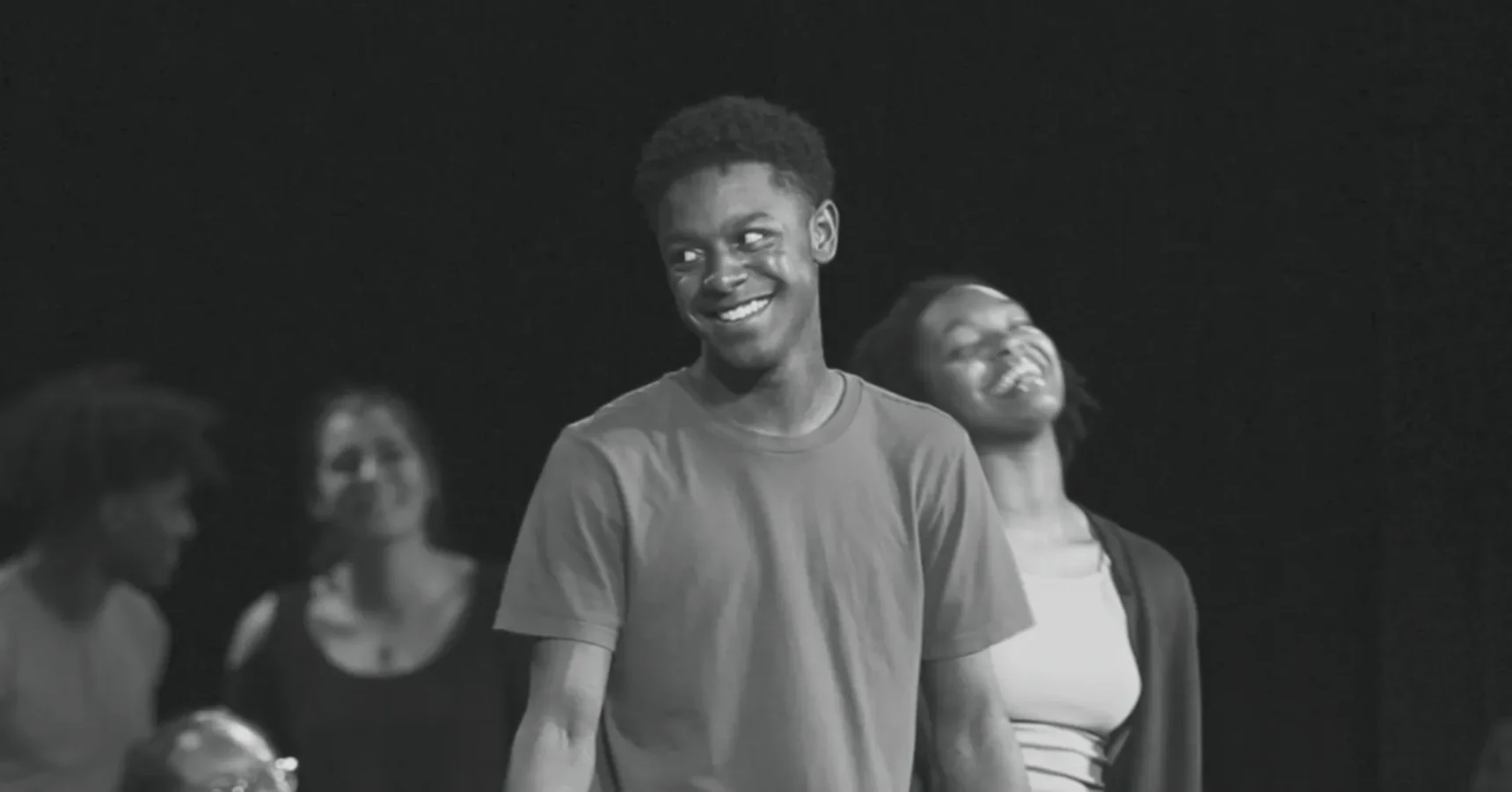 A young man in a t-shirt stands smiling, with a young woman behind him also smiling. Two other people are visible in the blurred background. The image is in black and white.