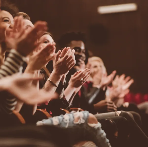 Audience members sitting in rows applaud, with hands mid-clap, in a dimly lit indoor setting.
