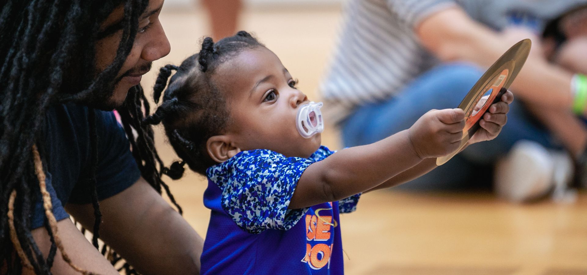 A baby with a pacifier holds up a circular object while sitting next to an adult on a wooden floor.