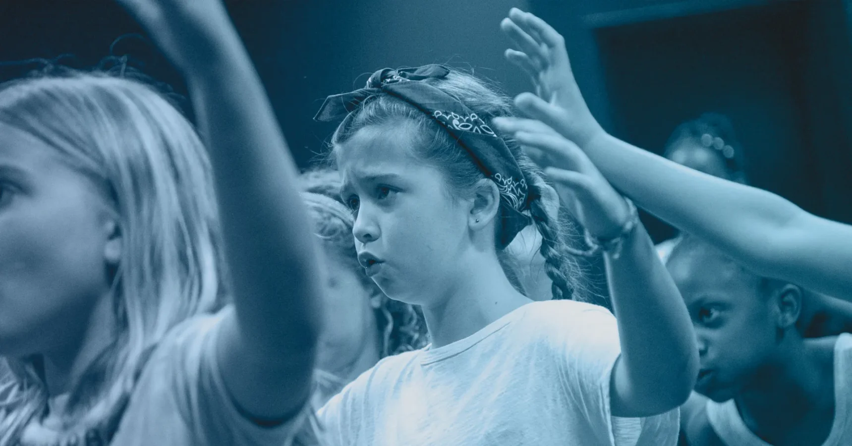 A group of children with raised arms participate in an activity; the focus is on a girl wearing a headband and a white shirt.