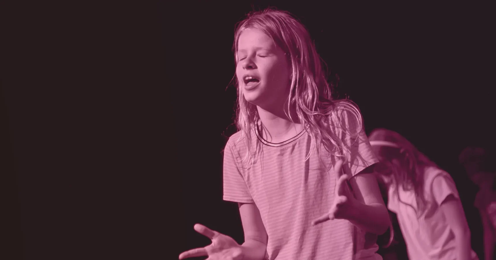 A young person stands with eyes closed and hands raised, appearing to be singing or speaking intensely on a dark stage.