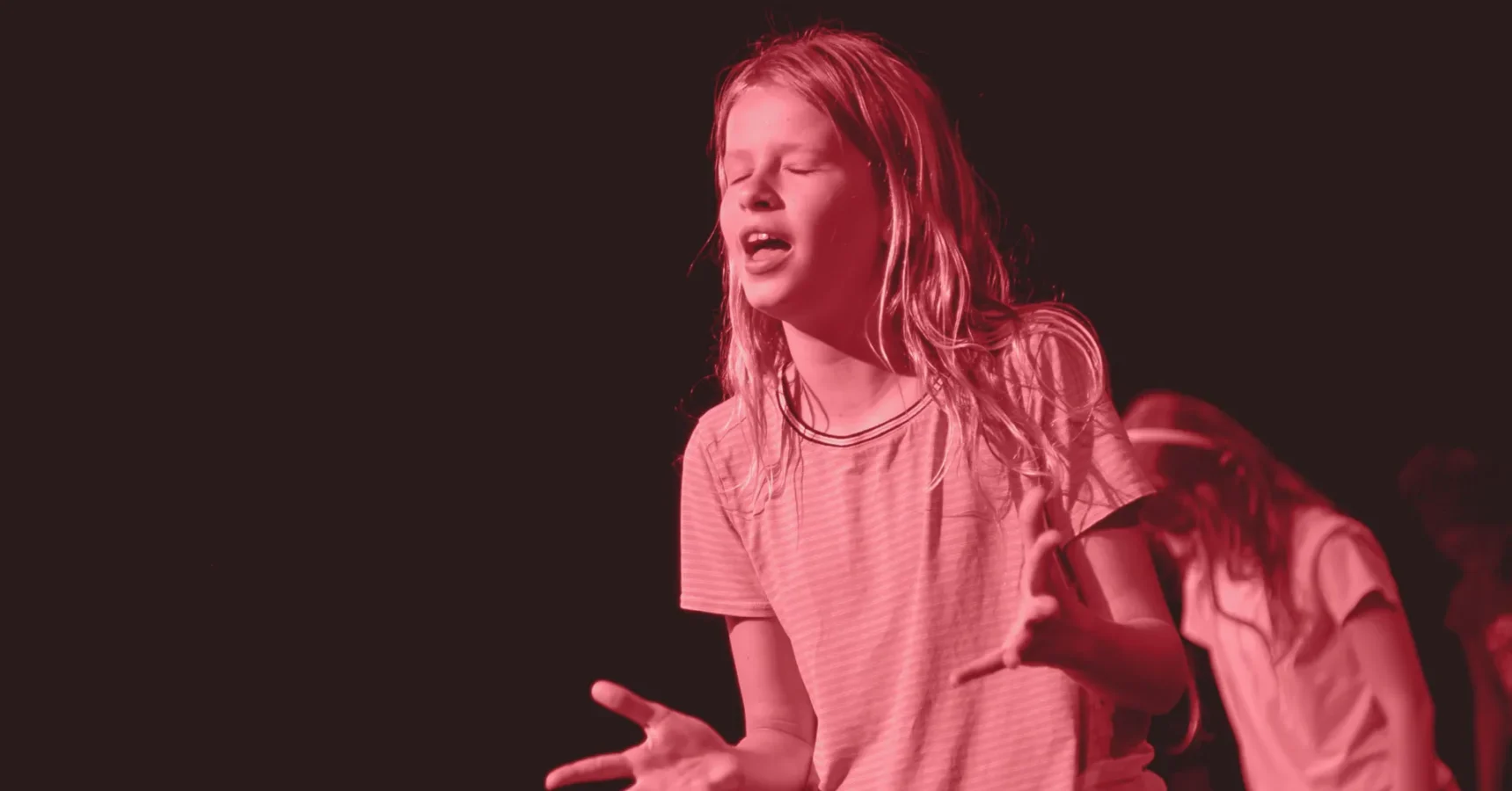 A young person with long hair stands with eyes closed and hands gesturing, appearing to be speaking or singing, against a dark background with red lighting.