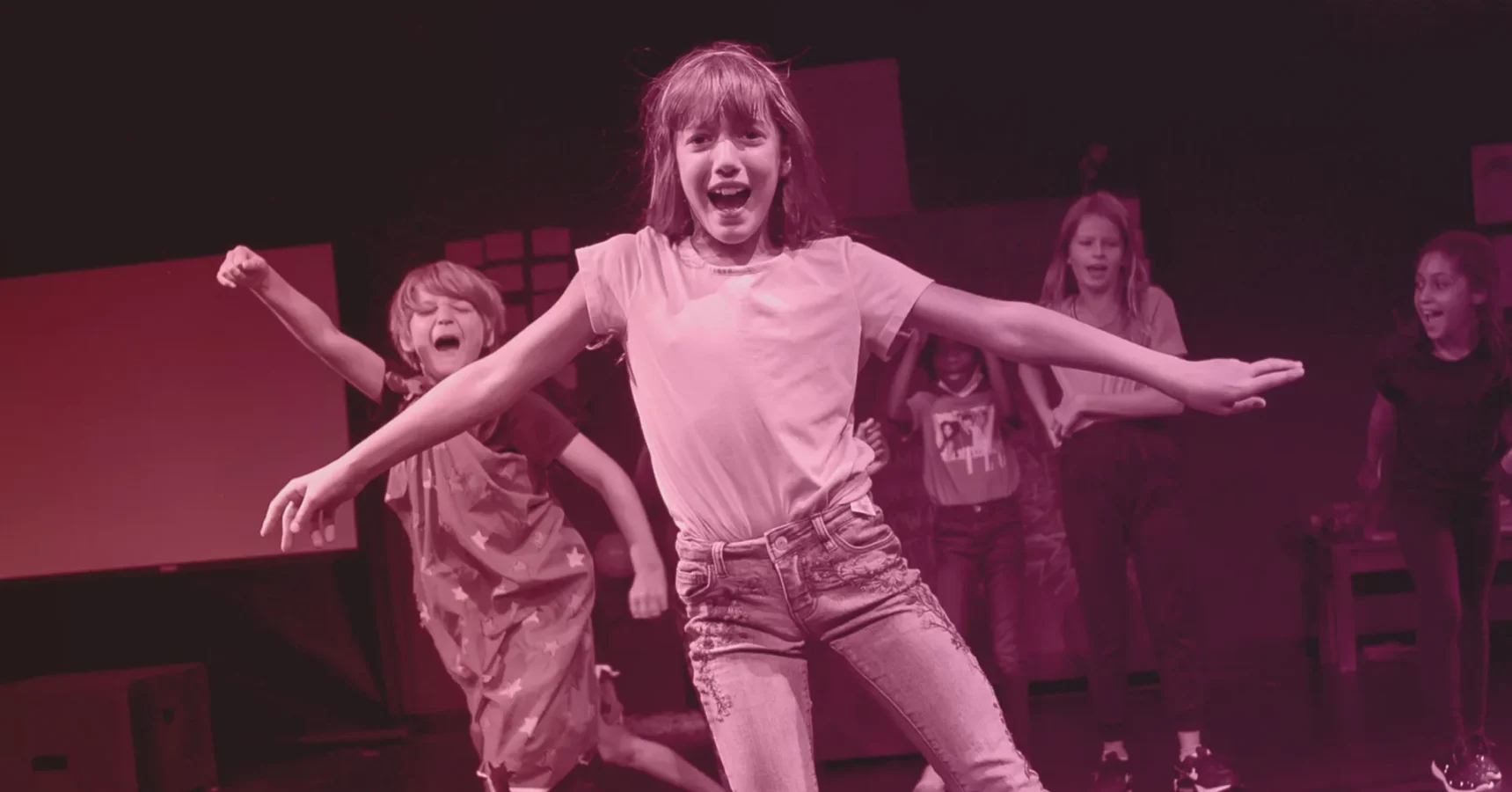Five children perform energetically on stage, with one girl in front spreading her arms while others cheer and dance behind her. The scene is tinted with a pink overlay.