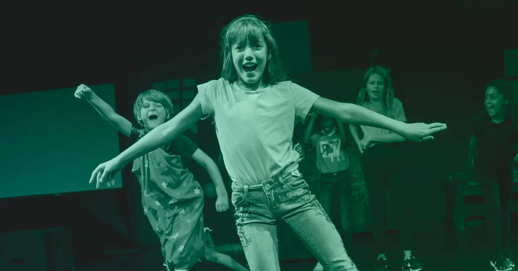 Several children are on stage, with one girl in the foreground posing with arms outstretched and smiling, while other children cheer and gesture behind her.