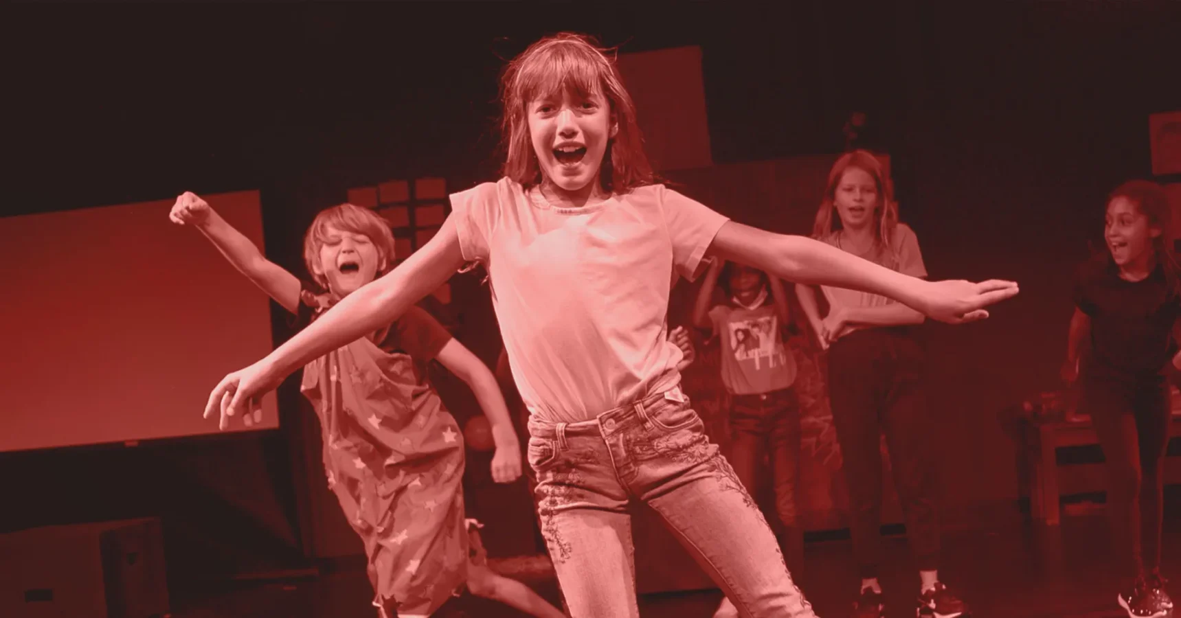 A group of children dance energetically on stage, with one girl in front leading the movement, all lit by red stage lighting.