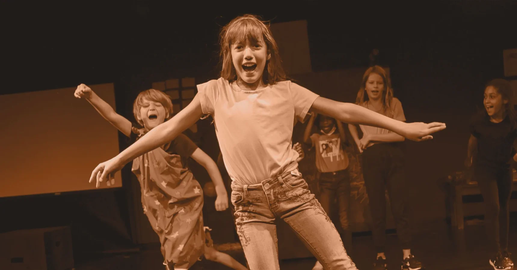 Five children perform energetically on stage, with one girl in front extending her arms and smiling, while the others pose and move in the background under stage lighting.