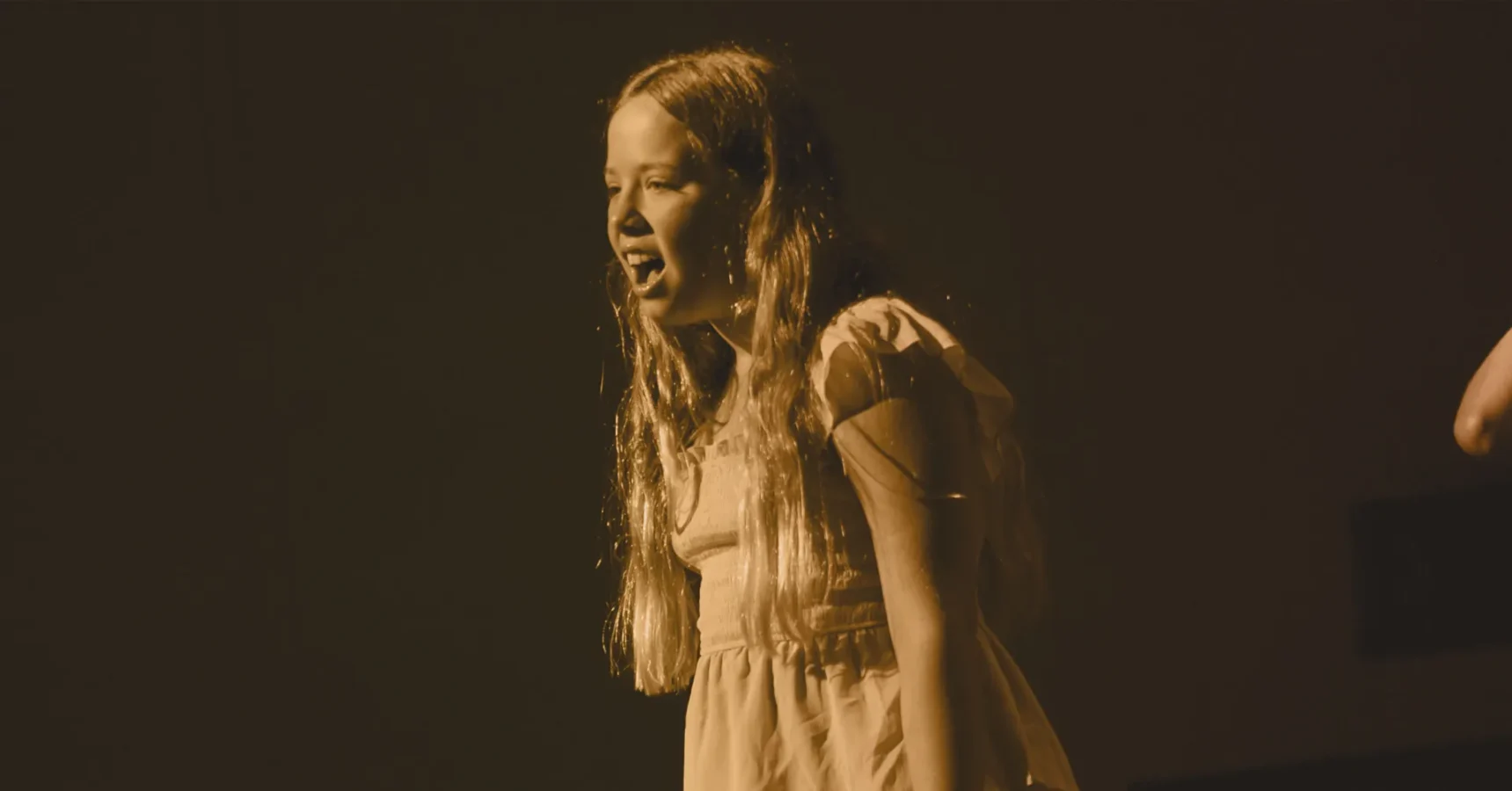 A young girl with long hair and a short-sleeve dress stands on stage, mid-speech or song, under a warm sepia light with a dark background.