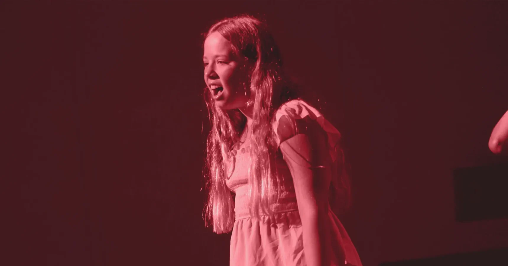 A girl with long hair stands on stage under red lighting, wearing a short-sleeved dress and appearing to sing or speak.