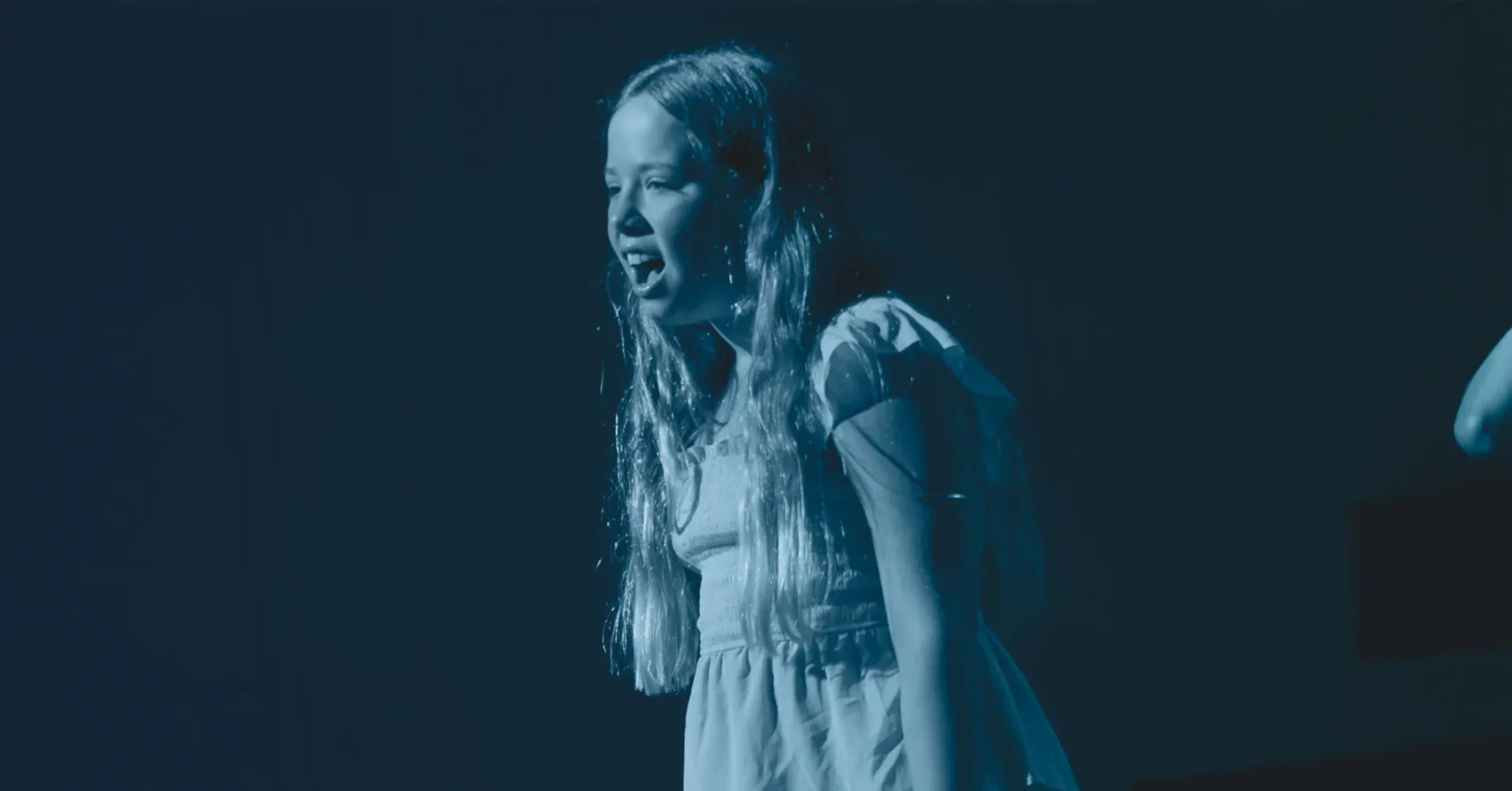 A young girl with long hair in a dress stands on stage, appearing to sing or speak, under blue lighting.