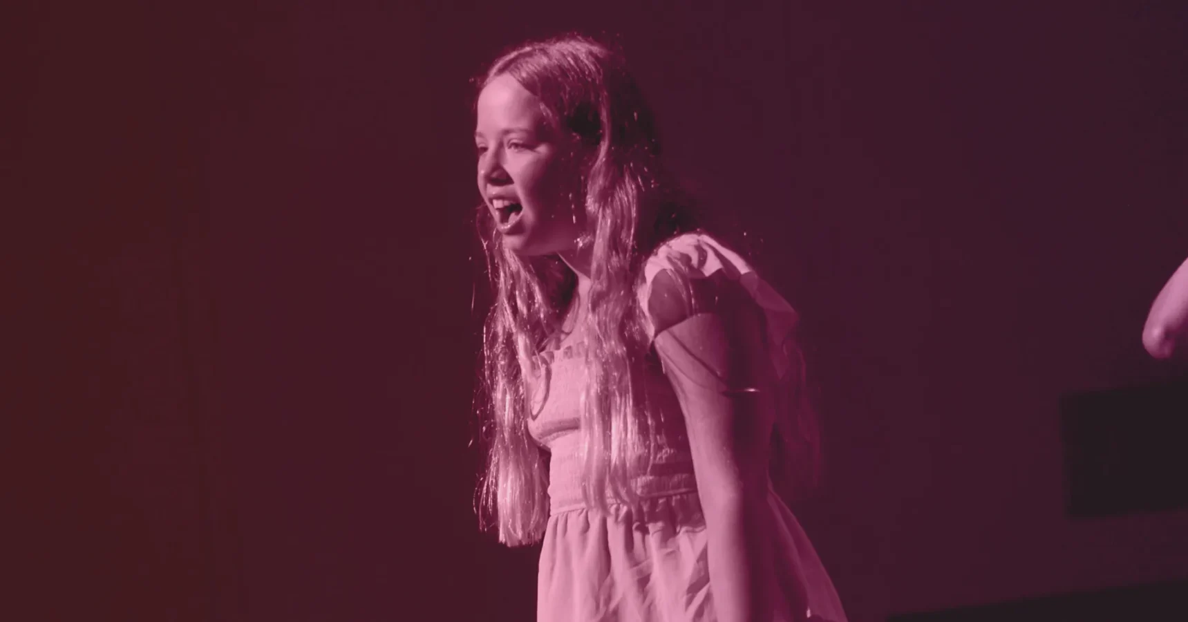 A young girl with long hair stands under stage lighting, wearing a short-sleeved dress and speaking or singing with an expressive face. The image has a pink color filter.