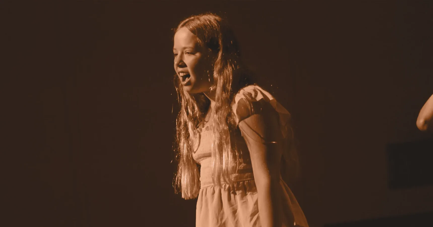 A young girl with long hair sings or speaks passionately on stage under warm lighting, wearing a short-sleeved dress.