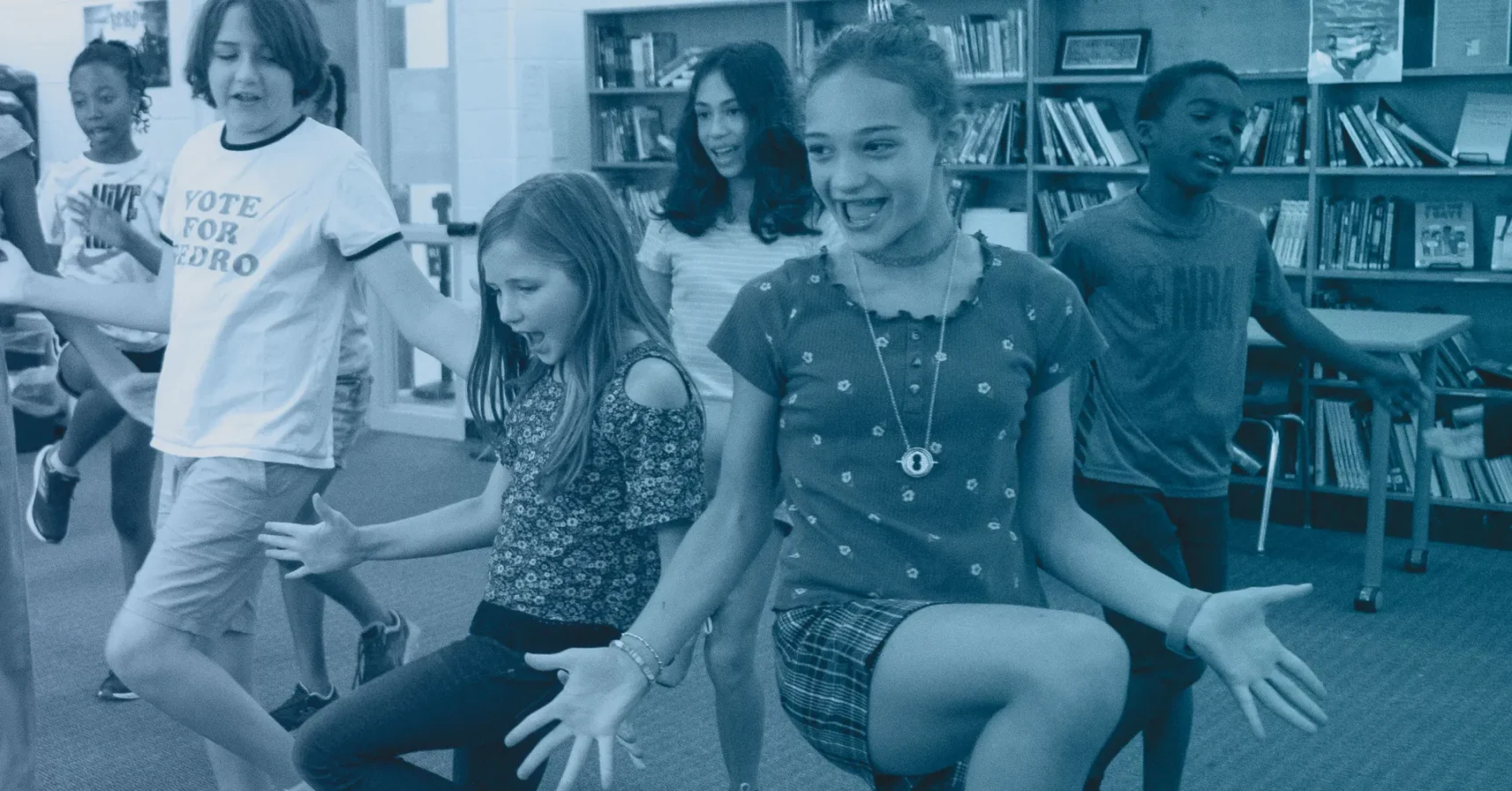 A group of children are dancing and smiling in a library, with bookshelves in the background.