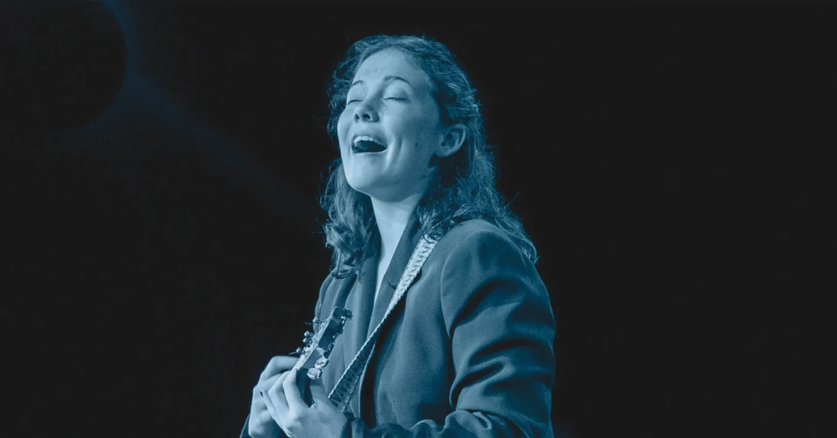 A person with curly hair plays guitar and sings on stage, wearing a jacket, against a dark background.