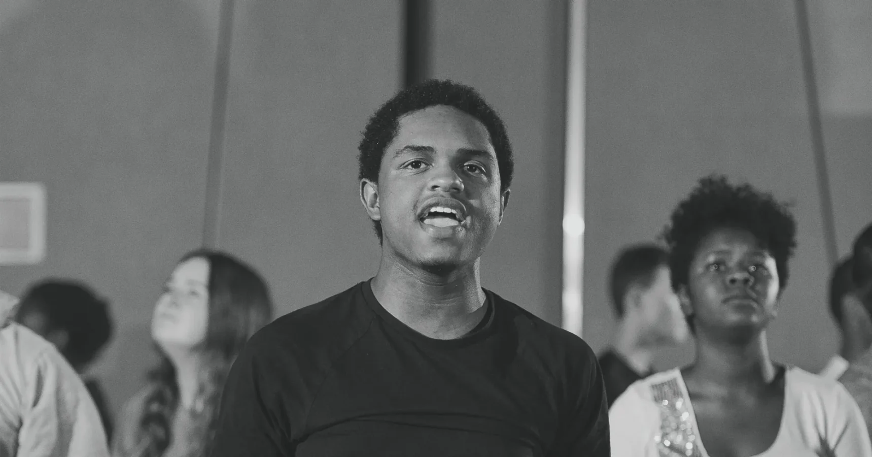 A group of people stand together indoors, with a young man in a dark shirt in the foreground speaking or singing. The image is in black and white.