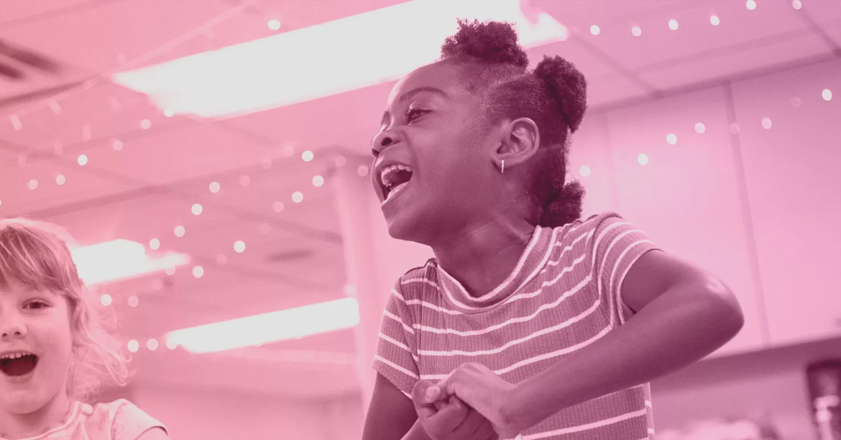Two children laughing and playing indoors under string lights, with a focus on a girl in a striped shirt smiling widely. The image has a pink color filter.