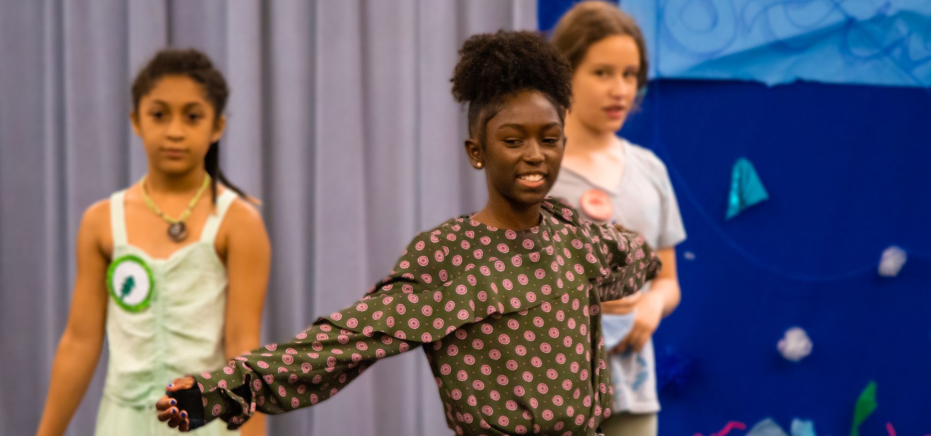 Three girls stand on stage; the girl in the foreground extends her arms and smiles, while two others stand behind her in front of a blue curtain and backdrop.