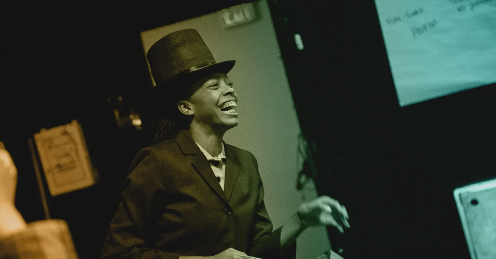 A person wearing a suit and tall hat smiles and gestures with one hand in a dimly lit indoor setting.