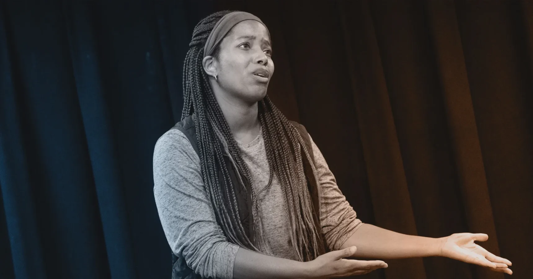 A person with long braids and a headband gestures with open arms while speaking, standing in front of a dark curtain.