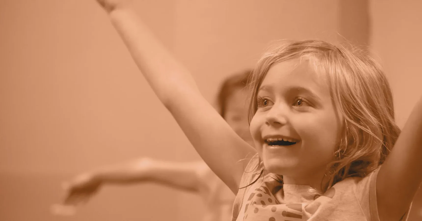 Smiling child with blonde hair raises both arms in excitement, with another child blurred in the background.