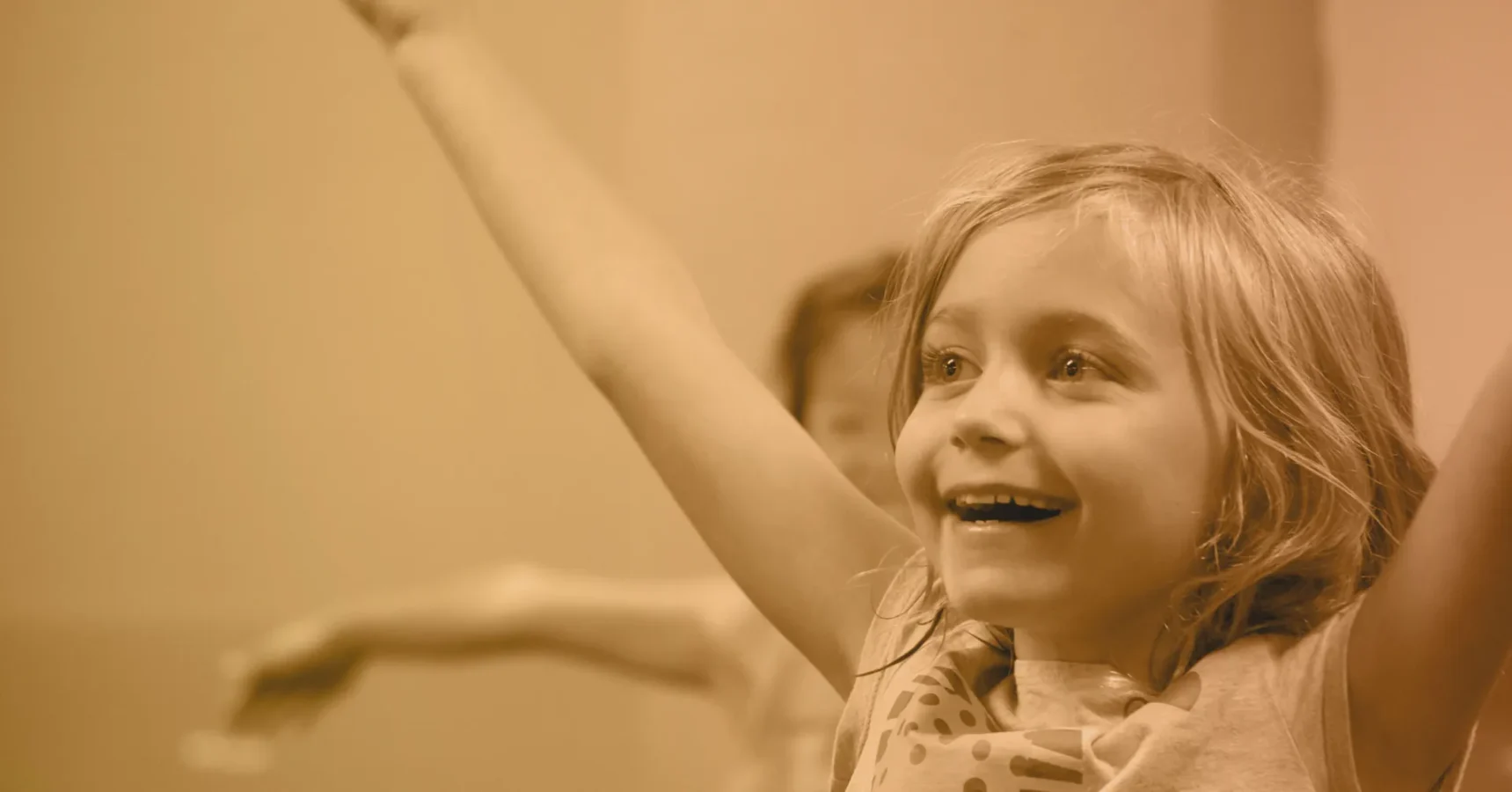 Smiling child with arms raised, looking upward, with another child blurred in the background.