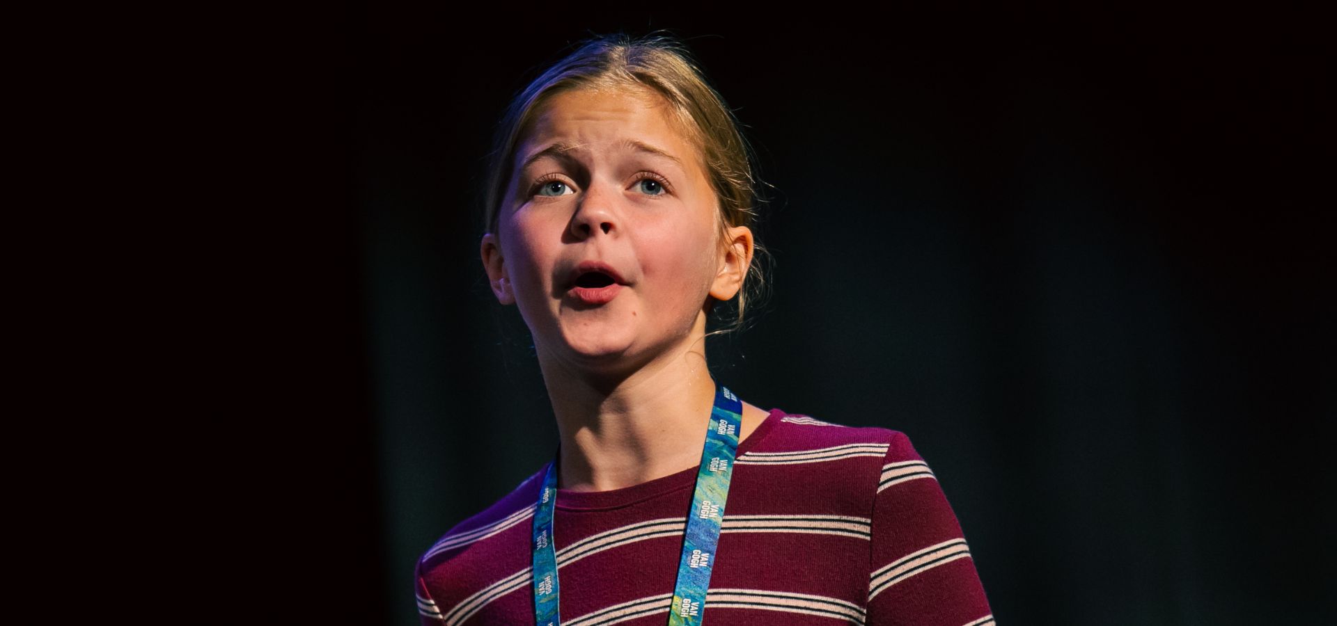A young girl with a striped shirt and event lanyard speaks on stage against a dark background.