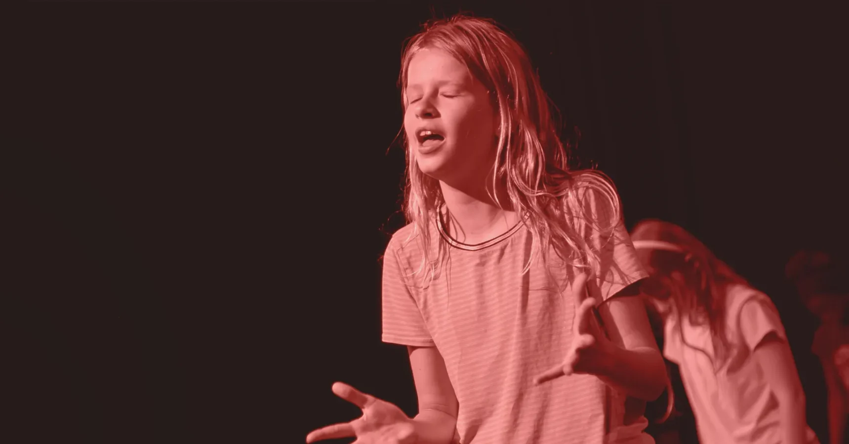 A young person with long hair and a striped shirt stands with eyes closed and mouth open, gesturing with both hands, under red stage lighting.