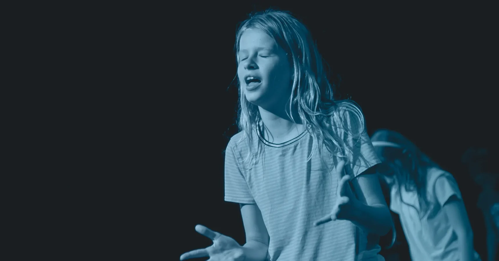 A young person with long hair stands with closed eyes and outstretched hands, appearing to sing or speak passionately against a dark background.