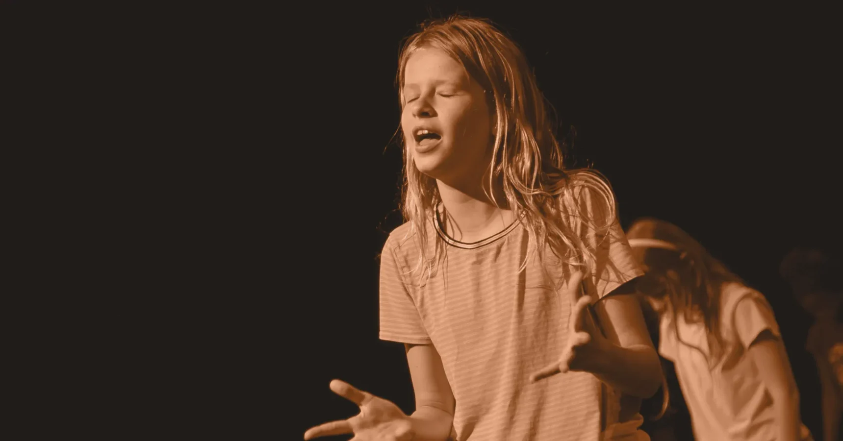 A young person with long hair and a striped shirt stands with eyes closed and hands raised, appearing to be performing or expressing emotion against a dark background.