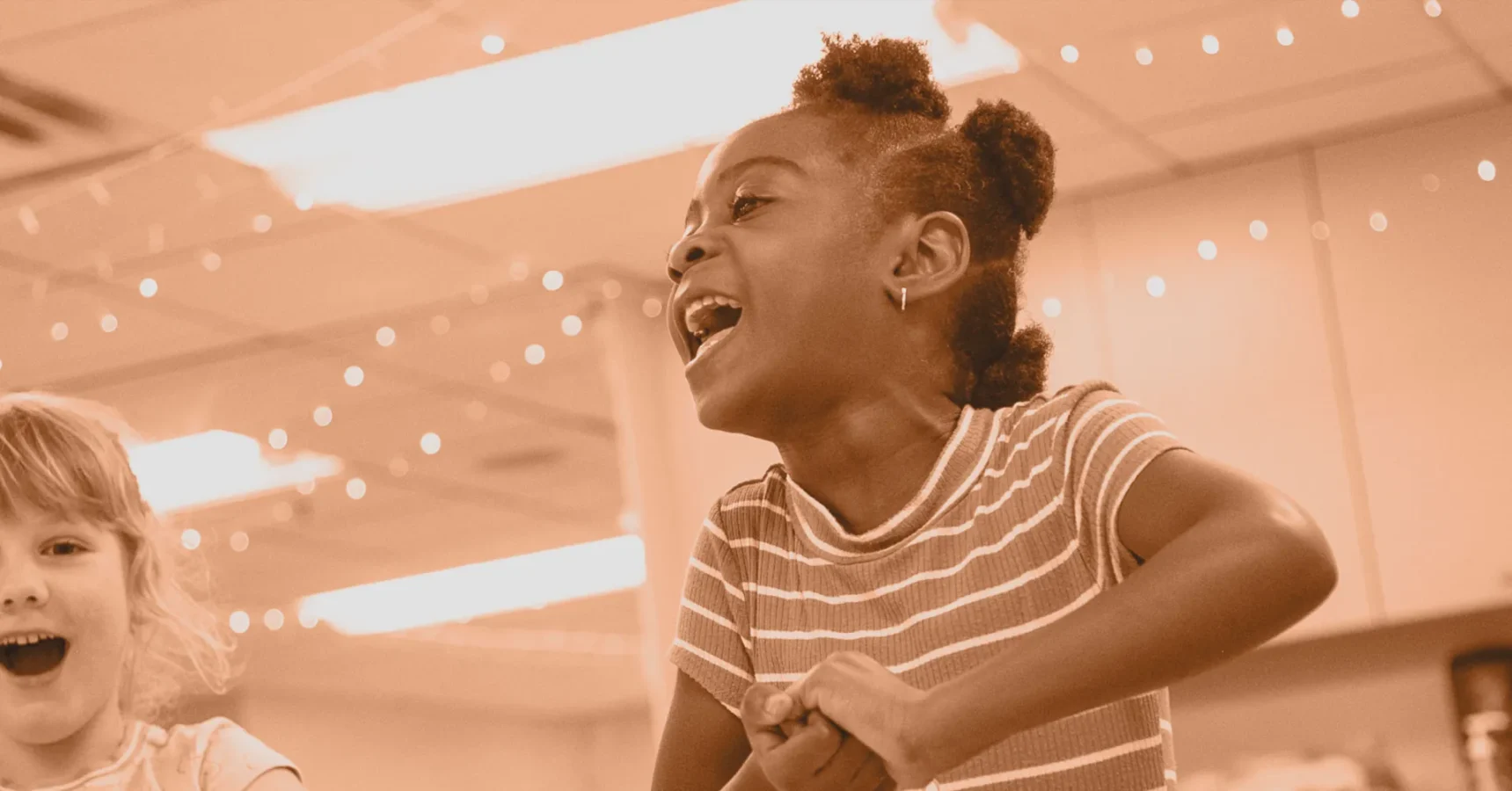 Two children indoors are smiling and laughing under string lights; one child in the foreground wears a striped shirt and looks excited.