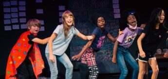 Five children stand in a line on stage, holding each other's shoulders and singing or performing in front of a chalkboard covered in writing and notes.