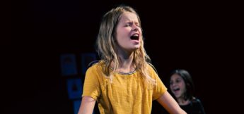 A young girl in a yellow shirt stands on stage with her mouth open, appearing to sing or perform, while another child is in the background at musical theater camp.