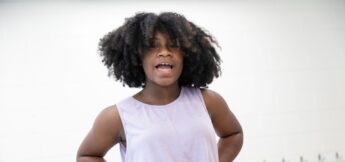 A young person with curly hair wearing a sleeveless lavender top stands with hands on hips, singing, against a white background in musical theater camp.