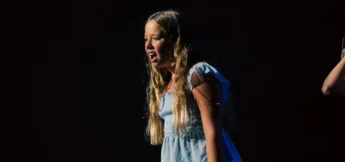 A young girl in a light blue dress stands on stage in musical theater camp under a spotlight, appearing to sing or speak with emotion against a dark background.