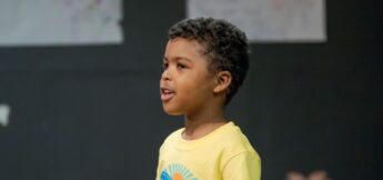 A young child wearing a yellow T-shirt stands indoors, singing, with a dark background and posters partially visible behind at musical theater camp.