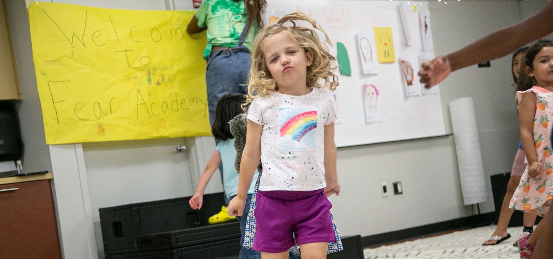 A young girl in a rainbow shirt and purple shorts poses confidently in a musical theater dance camp with other children and a sign in the background.