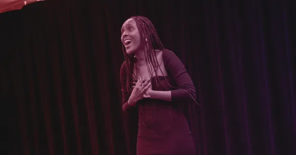 A woman in a long-sleeved dress stands on stage with her hands on her chest, speaking or performing in front of a black curtain.