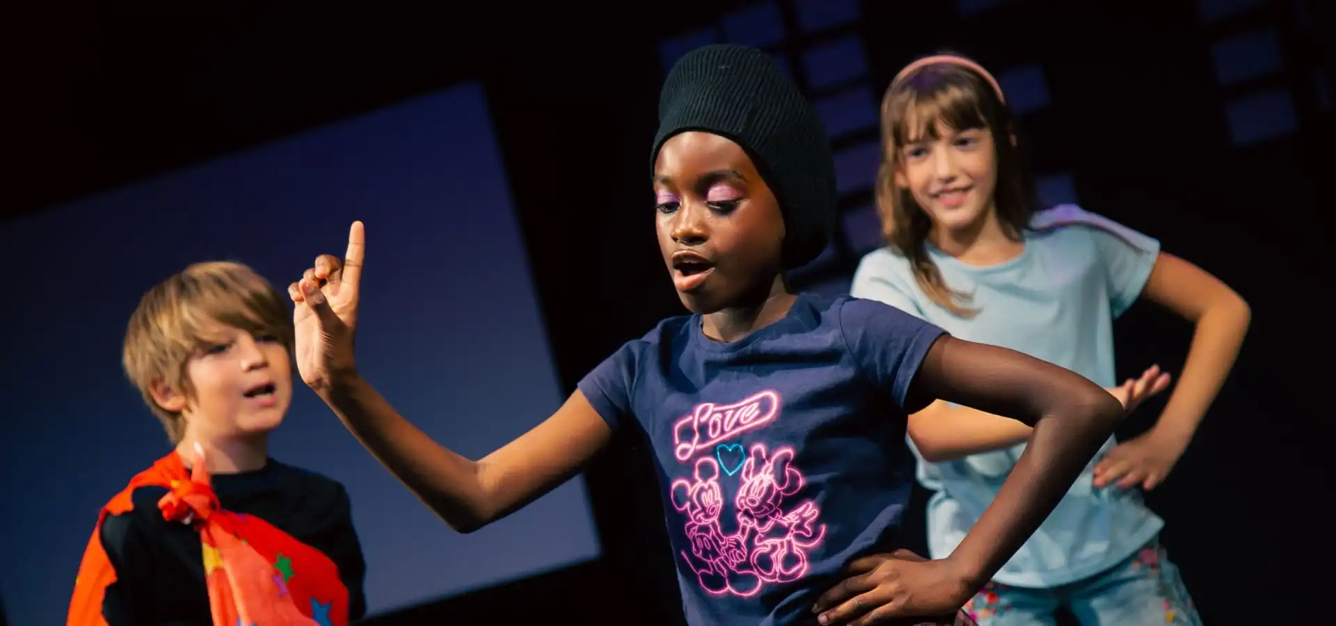 Three children perform on stage; the child in front gestures with one hand raised, while two others pose and smile in the background.