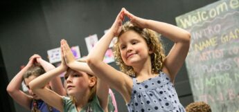 Children stand in a classroom with their hands pressed together above their heads, participating in an activity. A chalkboard with writing is visible in the background.