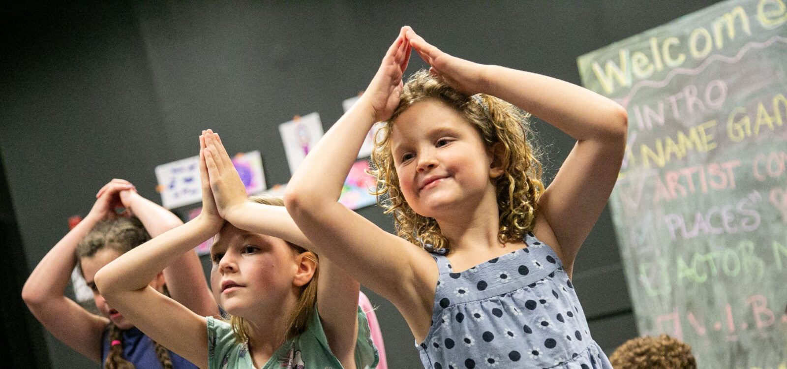 Children stand in a classroom with their hands pressed together above their heads, participating in an activity. A chalkboard with writing is visible in the background.