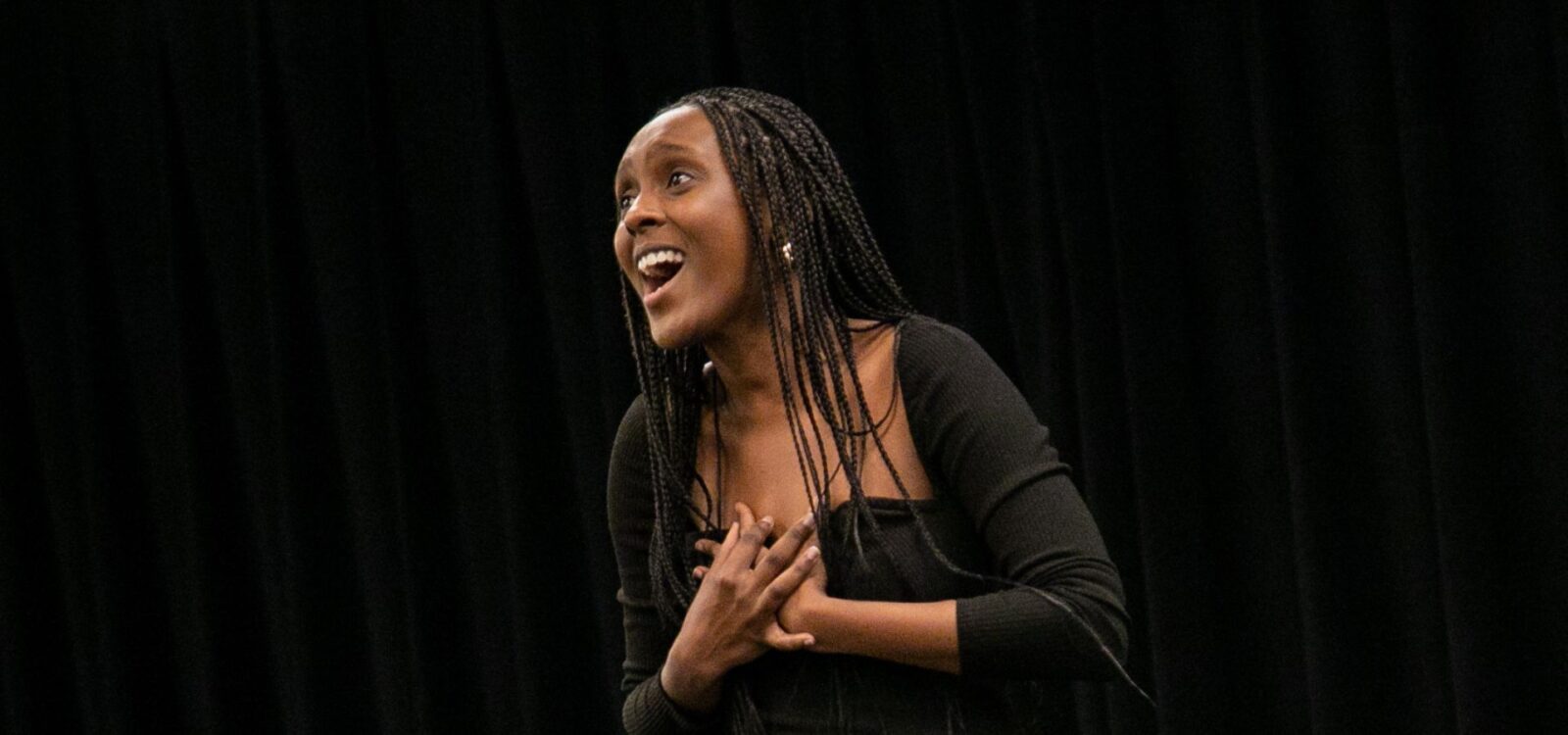 A woman with braided hair smiles and clasps her hands to her chest while standing in front of a black curtain.