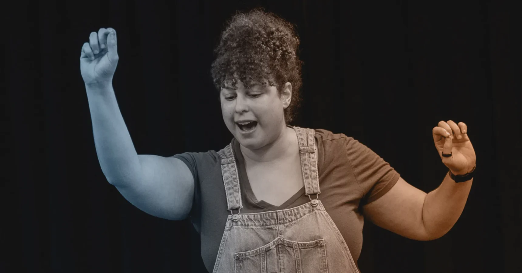 A person with curly hair, wearing overalls and a T-shirt, raises both arms while speaking or singing against a dark background.