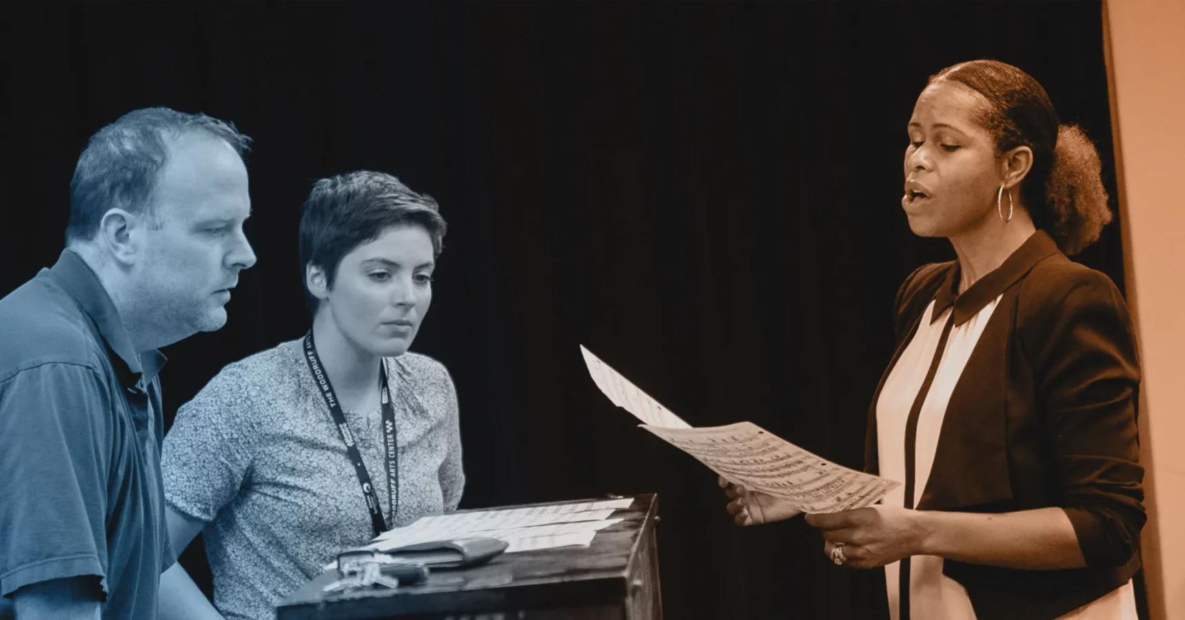 A woman stands and sings from sheet music while two people sit and observe, one man and one woman, in a dark indoor setting.