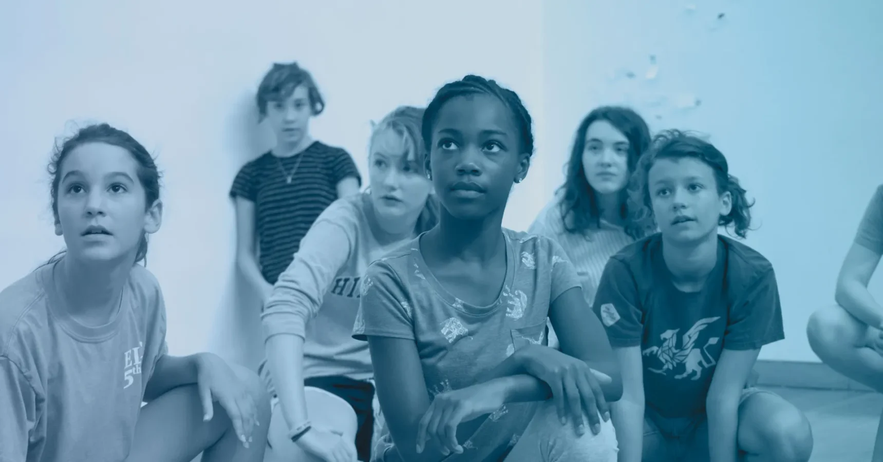 Six children sit on the floor indoors, looking attentively in the same direction. The photo uses a blue color filter.