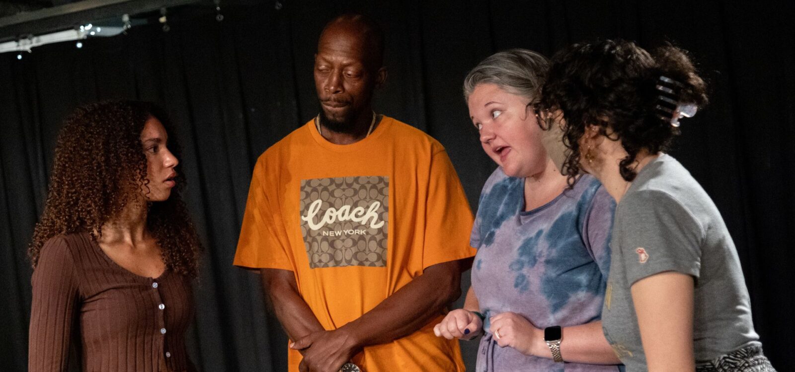 Four adults stand together indoors, engaged in a serious conversation against a black curtain backdrop.