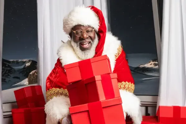 A man in a red and white Santa suit smiles while holding a stack of red gift boxes, with snowy mountains visible through the window—perfect for festive pictures with Santa.