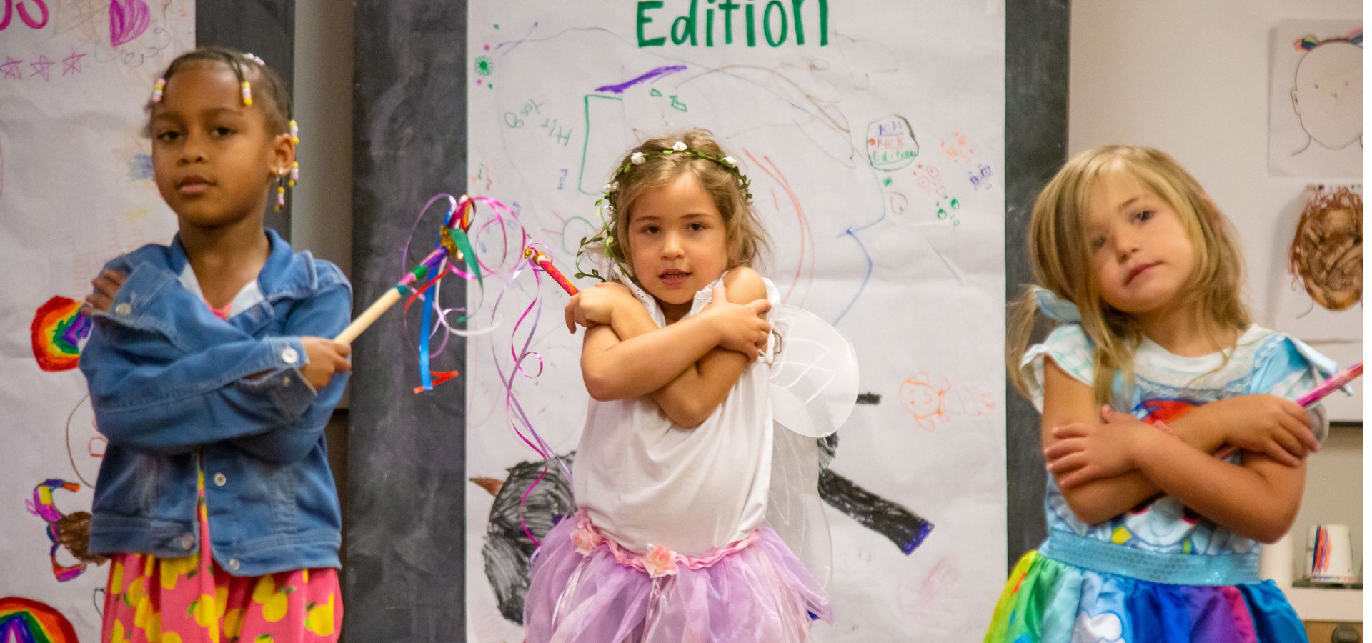 Three young girls stand in front of decorated posters, wearing colorful costumes and holding wands, with their arms crossed over their chests.