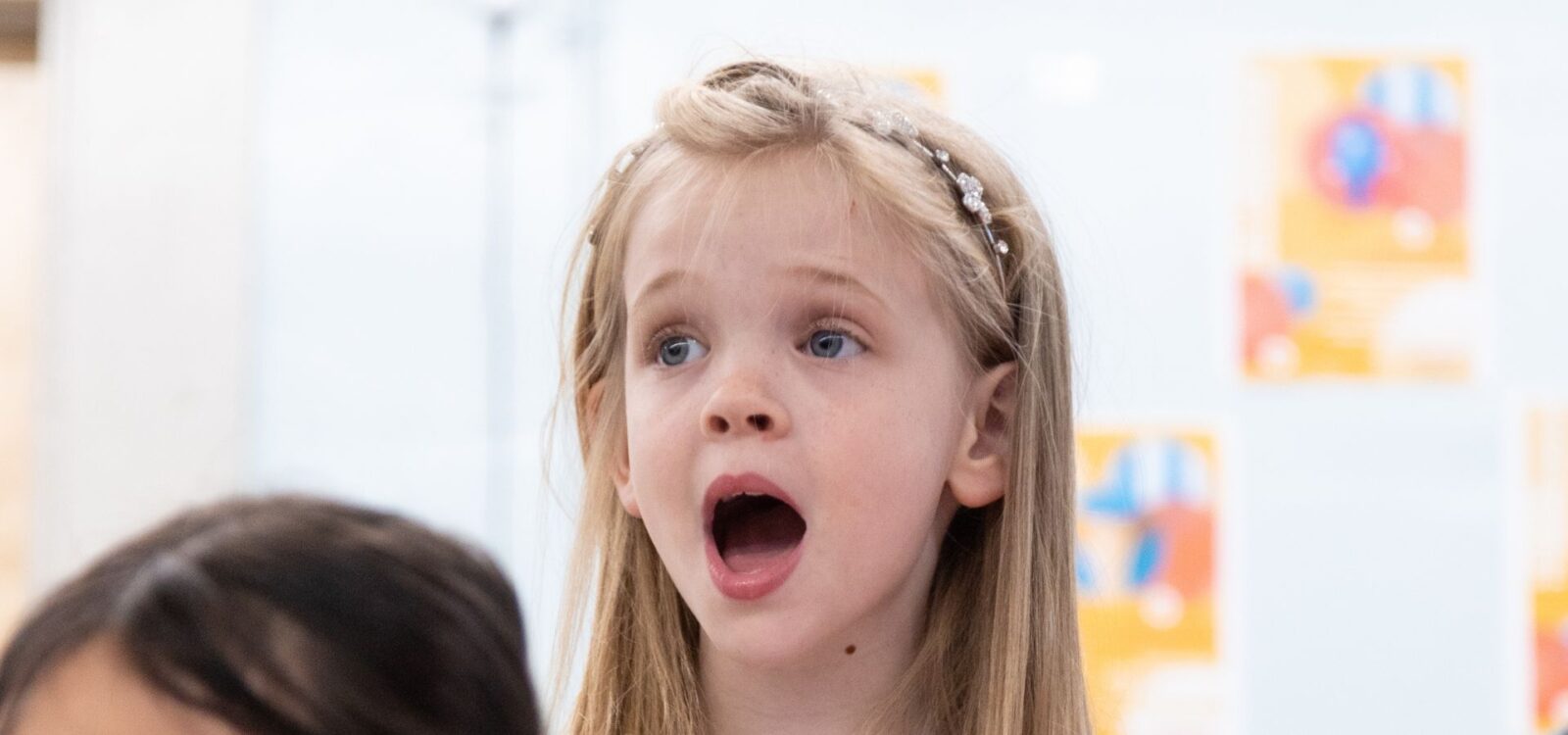 A young girl with long blonde hair and a pearl headband looks surprised with her mouth open. Colorful posters are blurred in the background.