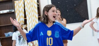 A girl wearing a blue Argentina soccer jersey with the number 10 stands indoors with her arms raised and smiling. Others are blurred in the background.